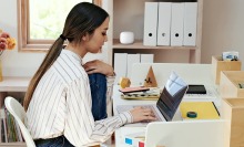 Woman working on laptop at desk with Google Nest mesh WiFi router on bookshelf behind her
