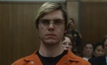 A man with glasses in an orange jumpsuit stands in a courtroom.