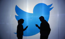 People are seen as silhouettes as they check mobile devices whilst standing against an illuminated wall bearing Twitter Inc.'s logo
