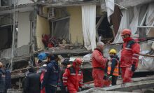 A group of emergency relief workers in red jumpsuits walk through the rubble of a building. 