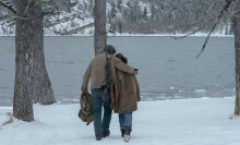 A man and a girl walk away from the camera through the snow, towards a lake.