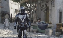 In a still from a sci-fi TV show, a man in full armour and helmet walks through a town with a small green creature travelling in a small spherical, hovering, vehicle.