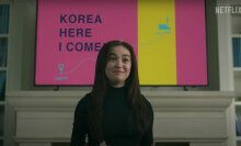 A young girl stands in front of a projector screen reading "Korea here I come".