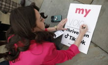 A woman in a pink blazer writes on a sign laid on the ground. The sign reads, "My voice thrives on TikTok."