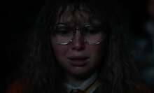 A woman with glasses stands in a dark room looking frightened.