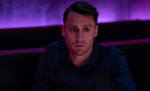 A man sits in a purple room looking troubled.