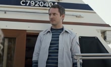 A man wearing a light blue collared shirt over a striped t-shirt looks suspicious standing on a boat.