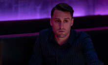 A man sits in a purple lit club room looking troubled.