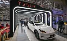 A white Tesla Model Y is parked in the glowing archway of a Tesla inspection bay. Employees work behind the vehicle.
