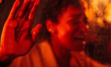 A woman presses her hand and face against a window and screams, bathed in red lighting.
