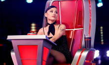 Ariana Grande sits in a large red and silver judges' chair on the set of The Voice. 