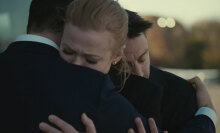 Three people in dark suits hug each other.