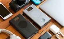 an array of devices are laid out on a wooden coffee table; the two power banks are in the center, surrounded by a camera, white headphones, an iphone, a silver laptop, and an airpods case 