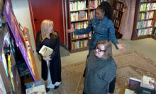 Two women stand in a bookstore, one looking at the shelves while another looks surprised by a third woman appearing behind them.