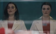 Two identical twin women in white lab coats and red shirts look down at a baby in an incubator.