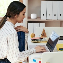 Woman working on laptop at desk with Google Nest mesh WiFi router on bookshelf behind her
