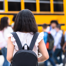 A girl wearing a backpack tentatively approaches a school bus with students waiting to board. 