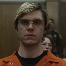 A man with glasses in an orange jumpsuit stands in a courtroom.