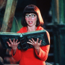 A woman in an orange sweater and black glasses holds a book and screams.