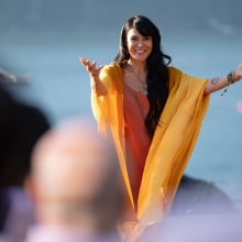 A woman in a yellow robe and orange dress holds her arms wide while addressing a crowd dressed in hues of purple.