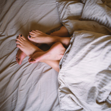 A couple lies in bed together, covered with a duvet while their legs are entwined.