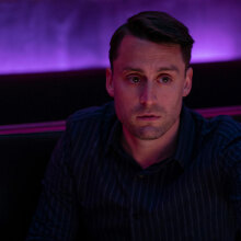 A man sits in a purple lit club room looking troubled.