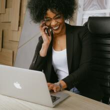 Business woman smiles on an iPhone while typing on a Mac laptop at a desk.