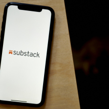 A phone sits on the edge of a wooden table as it's screen displays the Substack logo on a white background.