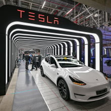 A white Tesla Model Y is parked in the glowing archway of a Tesla inspection bay. Employees work behind the vehicle.