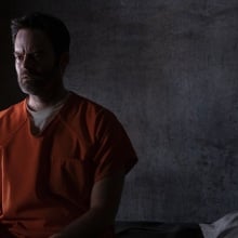 A man in an orange prison suit sits in a prison cell.