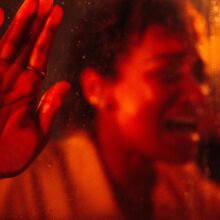 A woman presses her hand and face against a window and screams, bathed in red lighting.