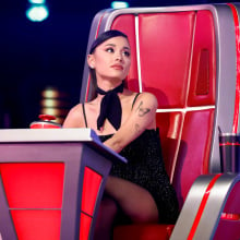 Ariana Grande sits in a large red and silver judges' chair on the set of The Voice. 