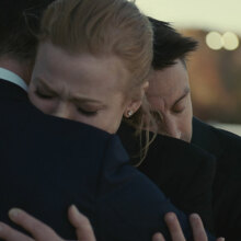 Three people in dark suits hug each other.
