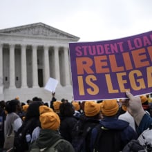 A crowd of protesters with yellow beanies stand in front of the U.S. Supreme Court. One person holds up a purple sign that reads, "Student loan relief is legal."