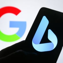 A silhouetted hand holds a phone with the blue Bing logo on the screen. Behind the phone is a Google logo in the background.