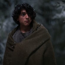 A young boy stands in the snow wearing a brown blanket. 