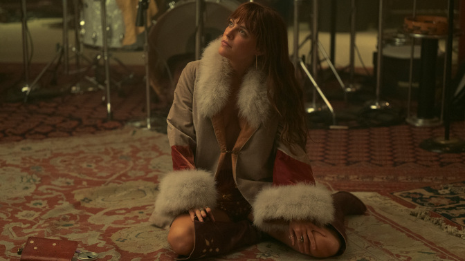 Daisy Jones sitting on the floor of the studio in a fur coat. 