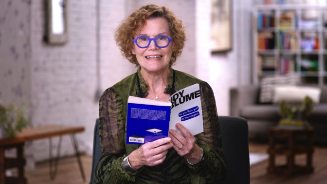 Judy Blume, wearing blue-rimmed glasses and a green blouse, reads from her book "Are You There God? It's Me, Margaret."