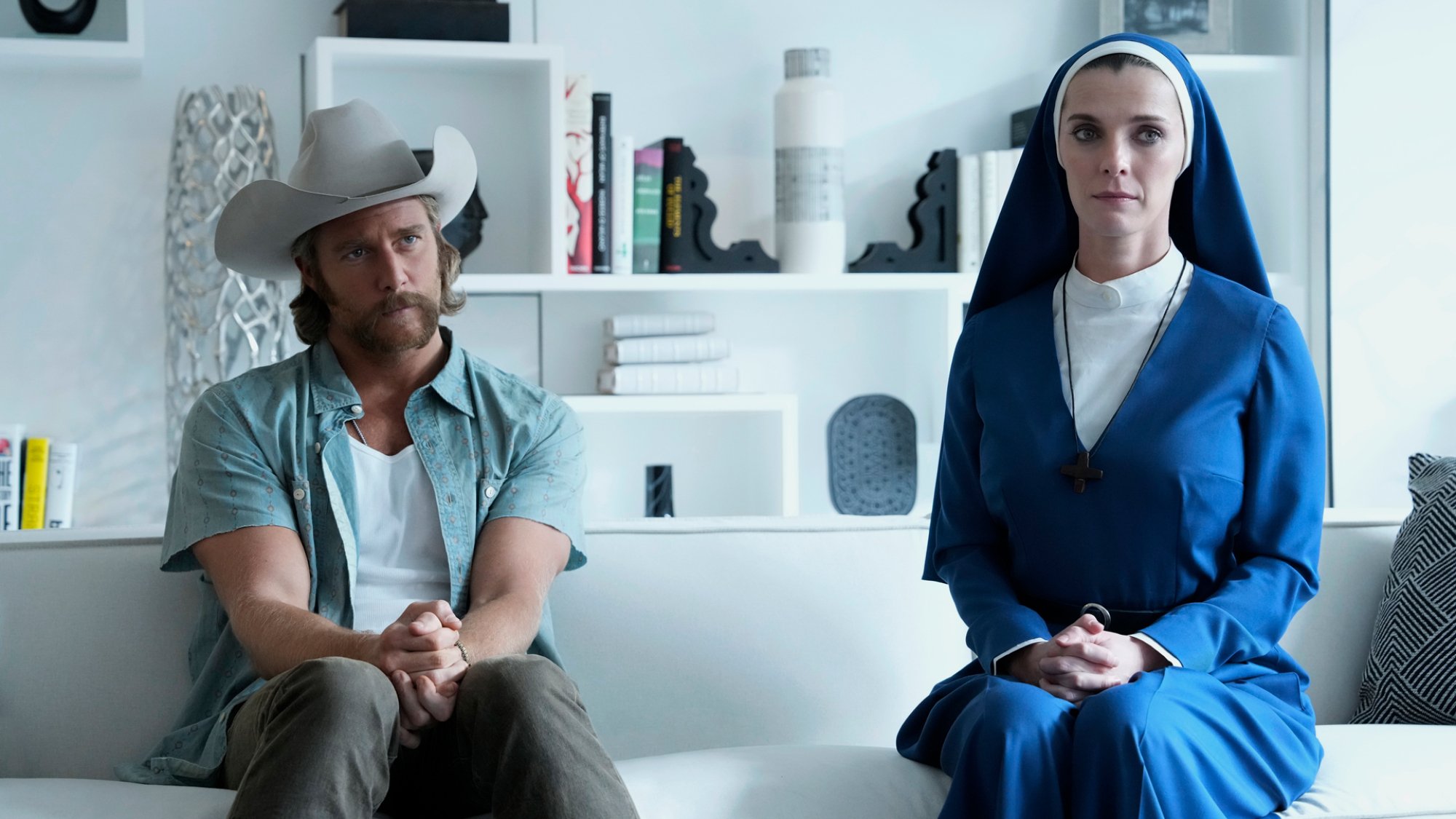 A man in a cowboy hat and a woman in a nun's outfit sit on a couch in a very white office.