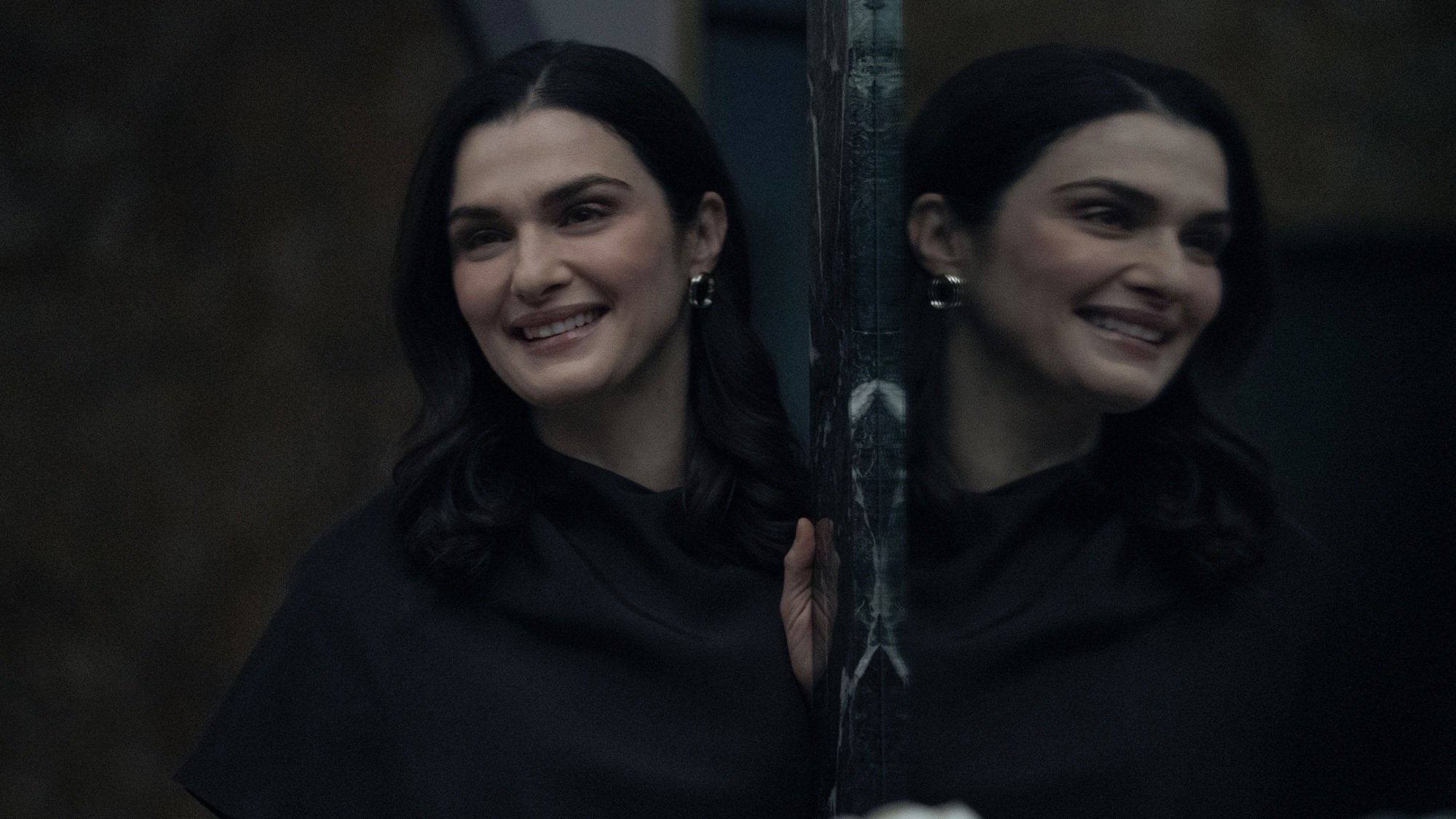 A smiling woman in a dark coat leans against a mirrored wall.
