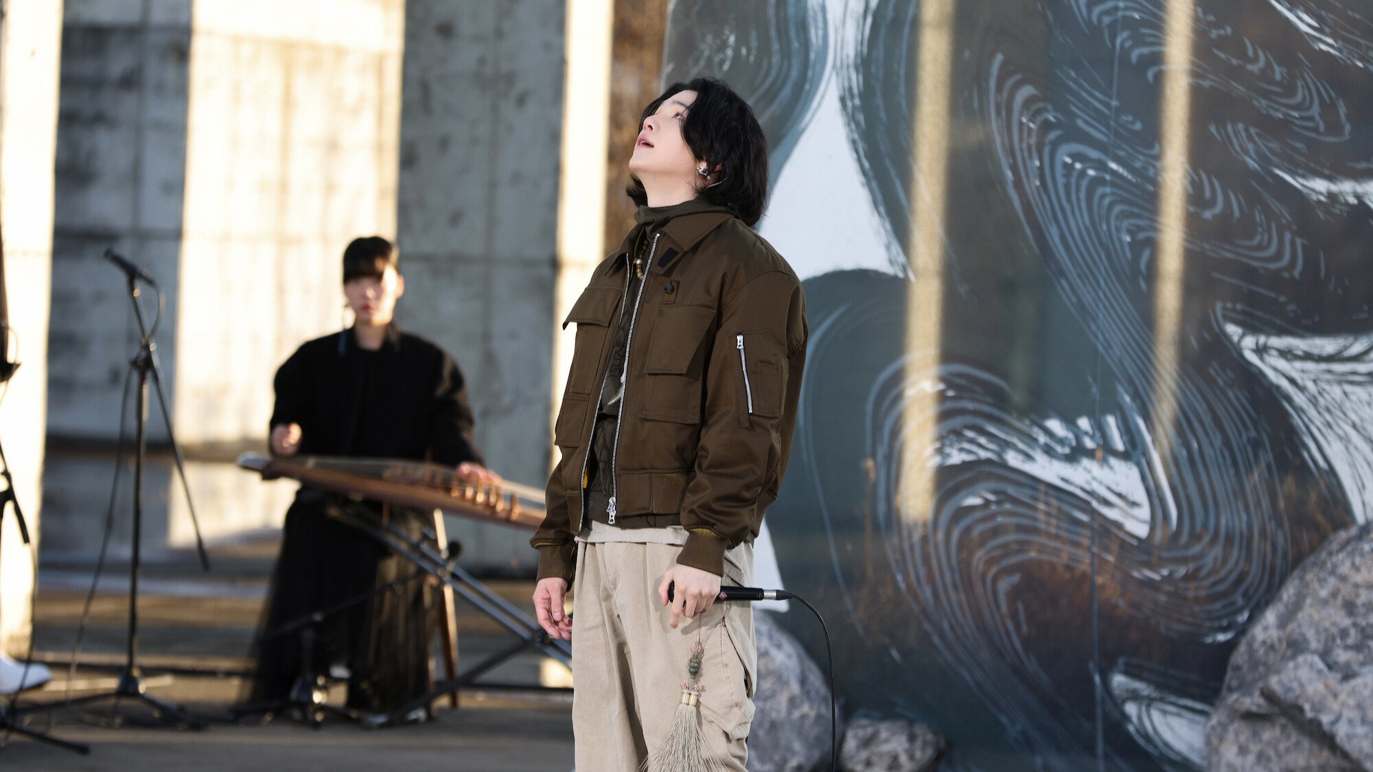 Suga from BTS performs outdoors in front of a black mural painting. 