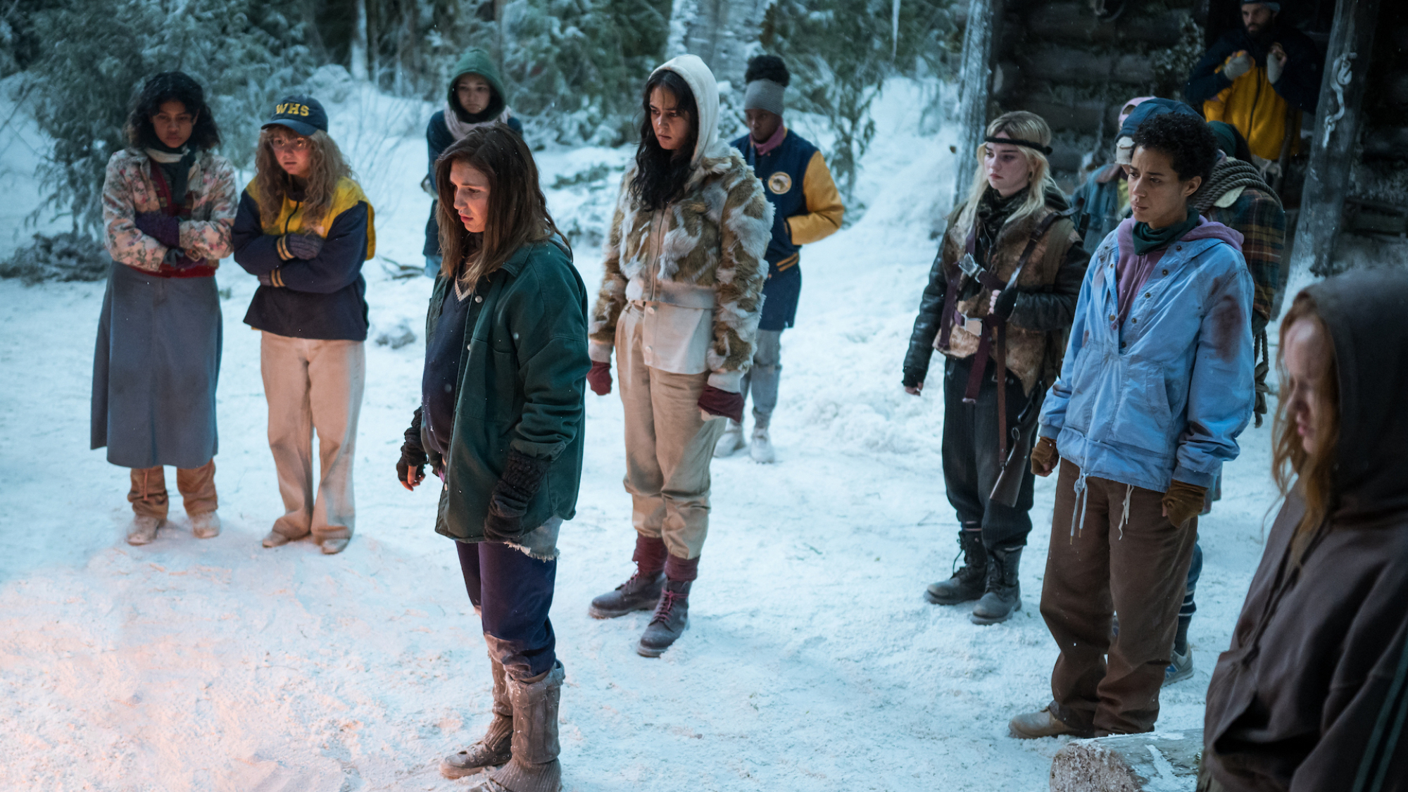A group of teen girls and two boys stand in the snow in vigil, as if at a funeral.