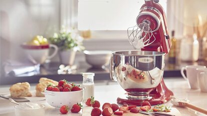 Red stand mixer with silver bowl and cream-covered whisk on table behind strawberries on chopping board and scones in background