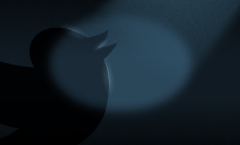 A darkly lit illustration of the Twitter logo bird in a black room. A very dim spotlight shines on its head. 