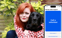 Lucy is crouching down and hugging a black Labrador in front of a garden wall. Next to her and the dog is a large iPhone screen showing the homepage of the Be My Eyes app, which is photoshopped into the picture. 