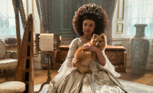 A young queen wearing a lavish dress, sits on a chair while holding a fluffy dog. 