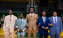 A group of teenage boys wearing suits stand next to each other with arms linked on a basketball court. 