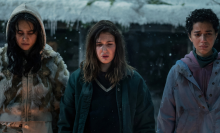 Three girls wearing thick coats stand in front of a fire while it snows. 