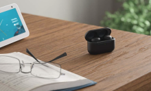 Black Amazon Echo Buds on a brown wood desk with a pair of glasses on top of a book on bottom left, tablet on upper left side