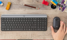 Black logitech keyboard, black logitech mouse on brown wooden desk with office supplies on the top